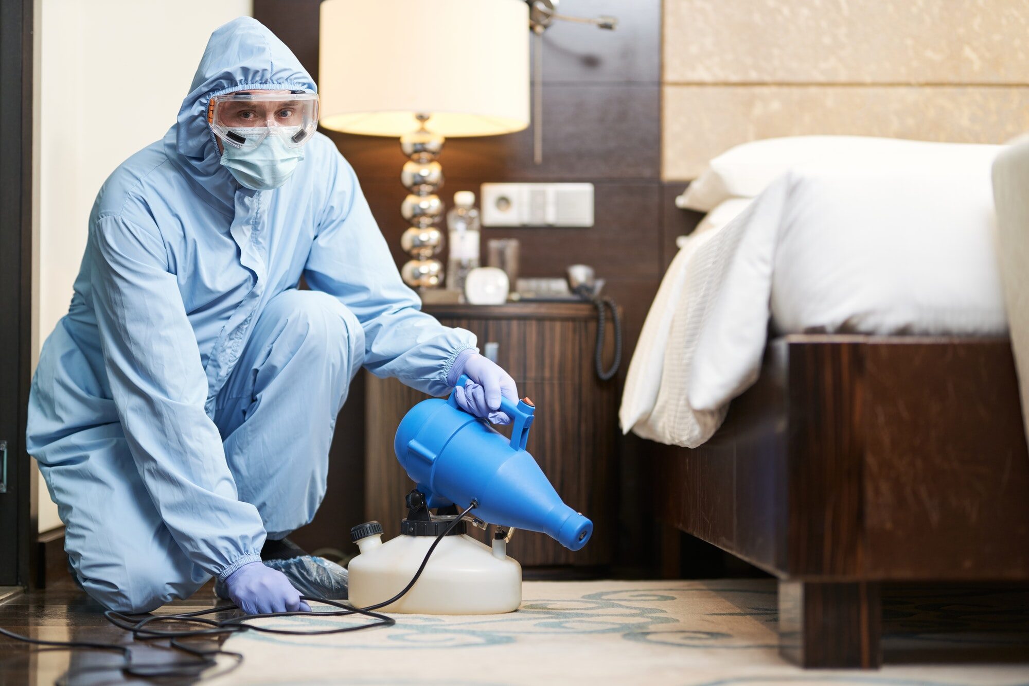 Worker in biohazard suit and mask disinfecting bedroom